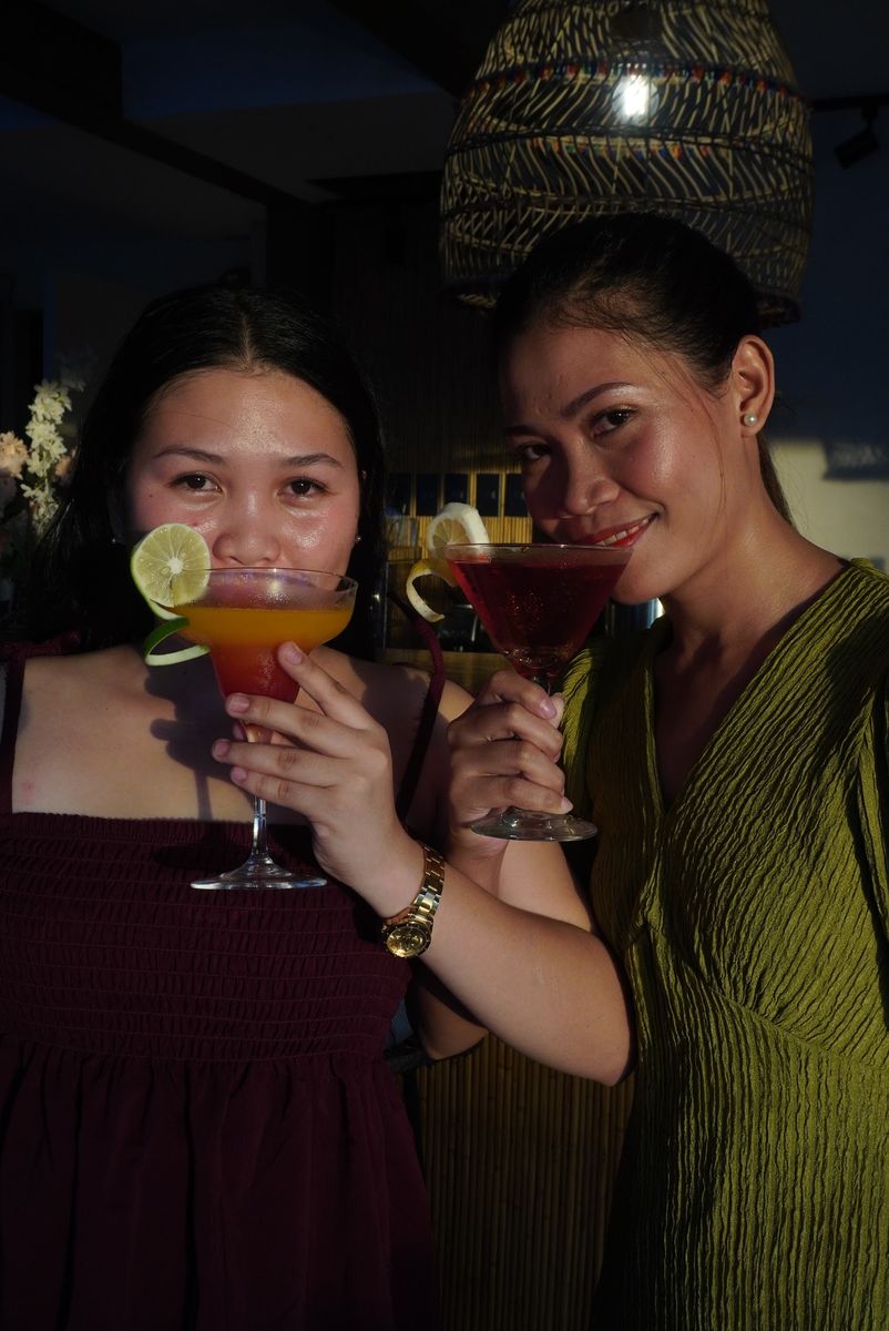 Two guests posing with signature cocktails under a woven rattan lamp.